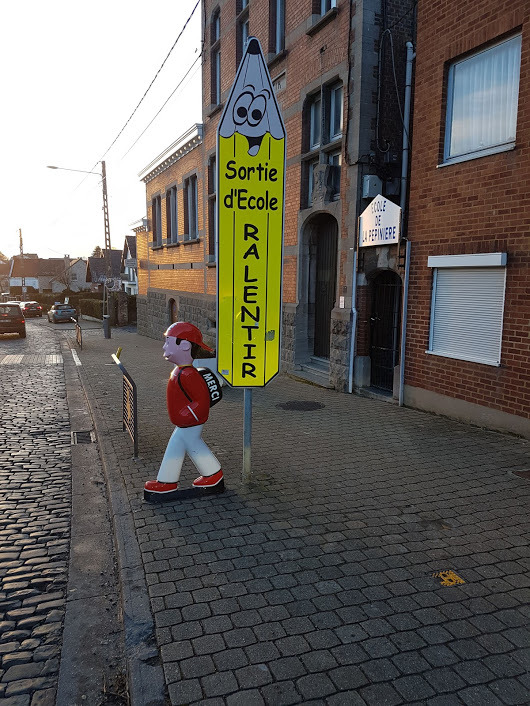 Żółty znak drogowy przed szkołą z napisem 'Sortie d'Ecole Ralentir' w kształcie ołówka z narysowaną twarzą, obok figura chłopca z napisem 'Merci' na plecaku, ustawione na chodniku z kostki brukowej.