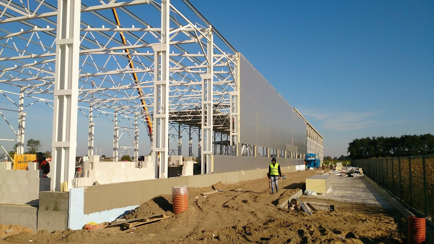 Wznoszenie konstrukcji stalowej hali: widok na częściowo obudowaną ścianę, biały szkielet konstrukcyjny, pracownik w żółtej kamizelce z łopatą na ziemi, w tle błękitne niebo.