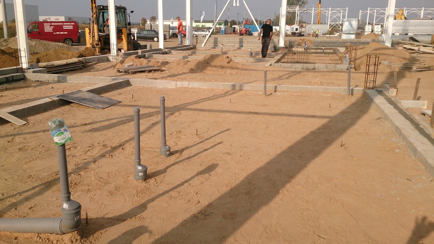 Fundamenty hali w trakcie budowy, widoczne zbrojenia, rury kanalizacyjne wystające z ziemi, piasek i sprzęt budowlany w tle.