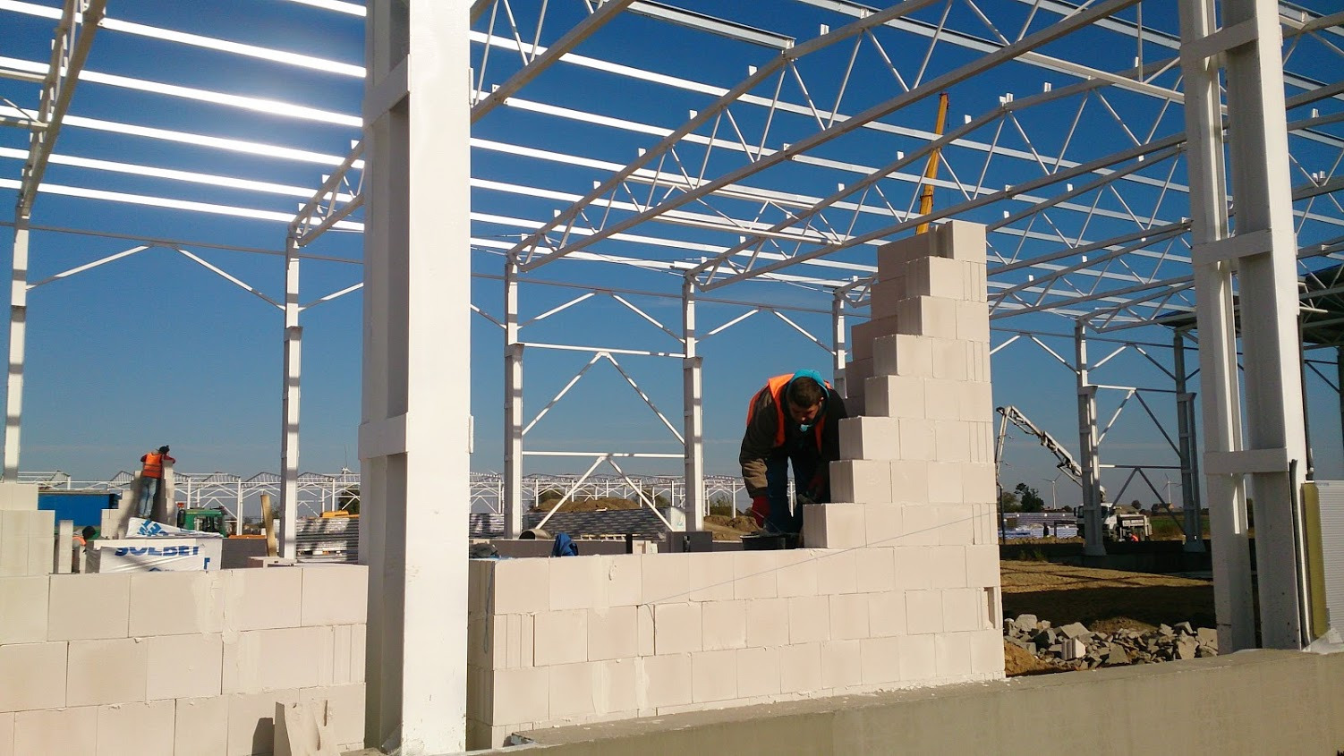Wznoszenie ściany z białych bloczków na tle konstrukcji stalowej hali, pracownik w kamizelce i masce murarskiej układa kolejne warstwy, na zewnątrz widoczny krajobraz i maszyny budowlane.