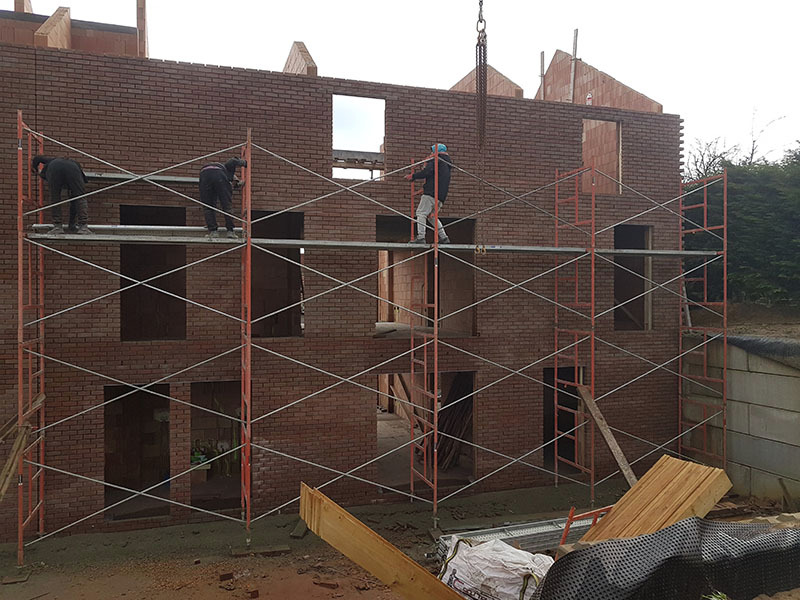 Murarze pracują na rusztowaniu przy wznoszeniu dwukondygnacyjnego budynku z cegły klinkierowej, widoczne niezamurowane otwory okienne i drzwiowe, oraz dźwig podający materiał.