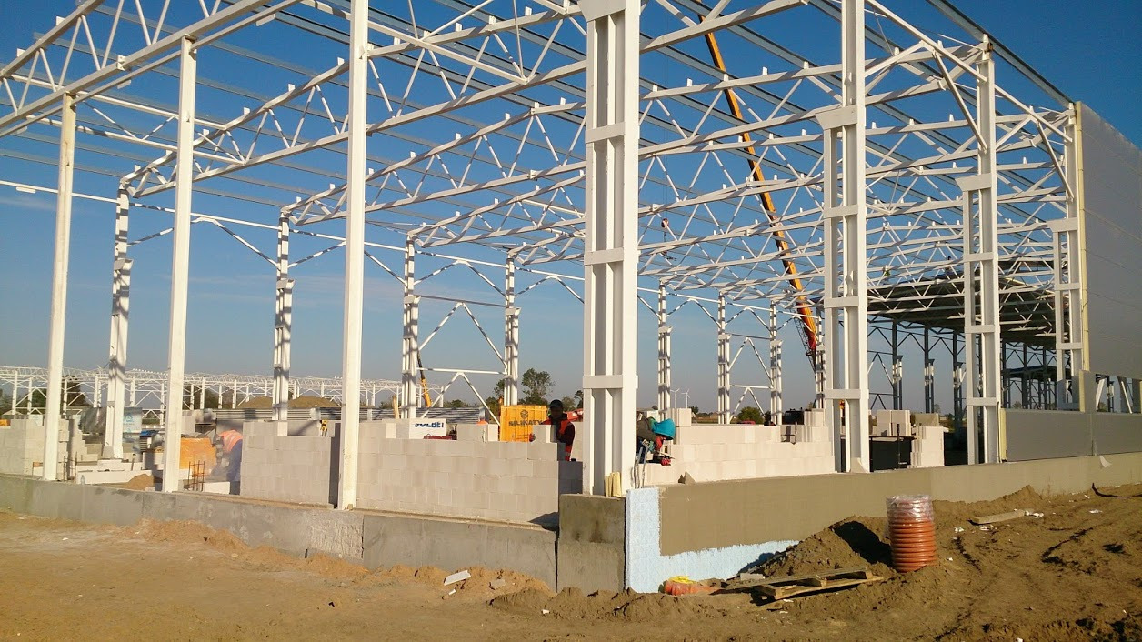 Wznoszenie szkieletu hali stalowej: widoczne białe konstrukcje nośne, murowane ściany z bloczków, ocieplenie fundamentów styropianem, robotnicy w odblaskowych ubraniach oraz dźwig w tle.