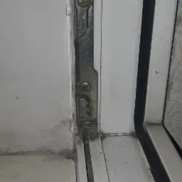 Uszkodzone okucie okienne z widocznymi śladami korozji i pęknięciem, przymocowane dwoma śrubami do białej ramy okiennej.