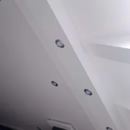 Biały sufit podwieszany z oświetleniem punktowym LED, widok pod kątem, z fragmentem ciemnej ściany i rośliny w tle.