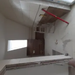 Montaż białych płytek na ścianie w łazience pod skosem, widoczna poziomica, drabina i okno dachowe.