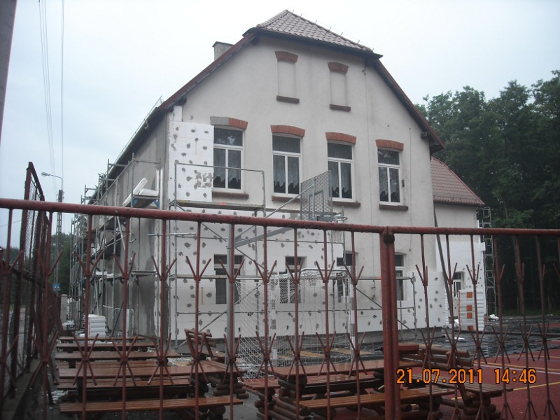 Budynek szkolny w trakcie docieplania, widoczne rusztowania i płyty styropianowe z resztkami kleju, czerwona metalowa bariera na pierwszym planie i data wykonania zdjęcia w prawym dolnym rogu.