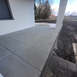 Nowoczesny taras z szarych płyt betonowych, zarys budynku w tle, ziemia przygotowana pod nasadzenia. Minimalistyczny design z widocznymi detalami wykończenia.