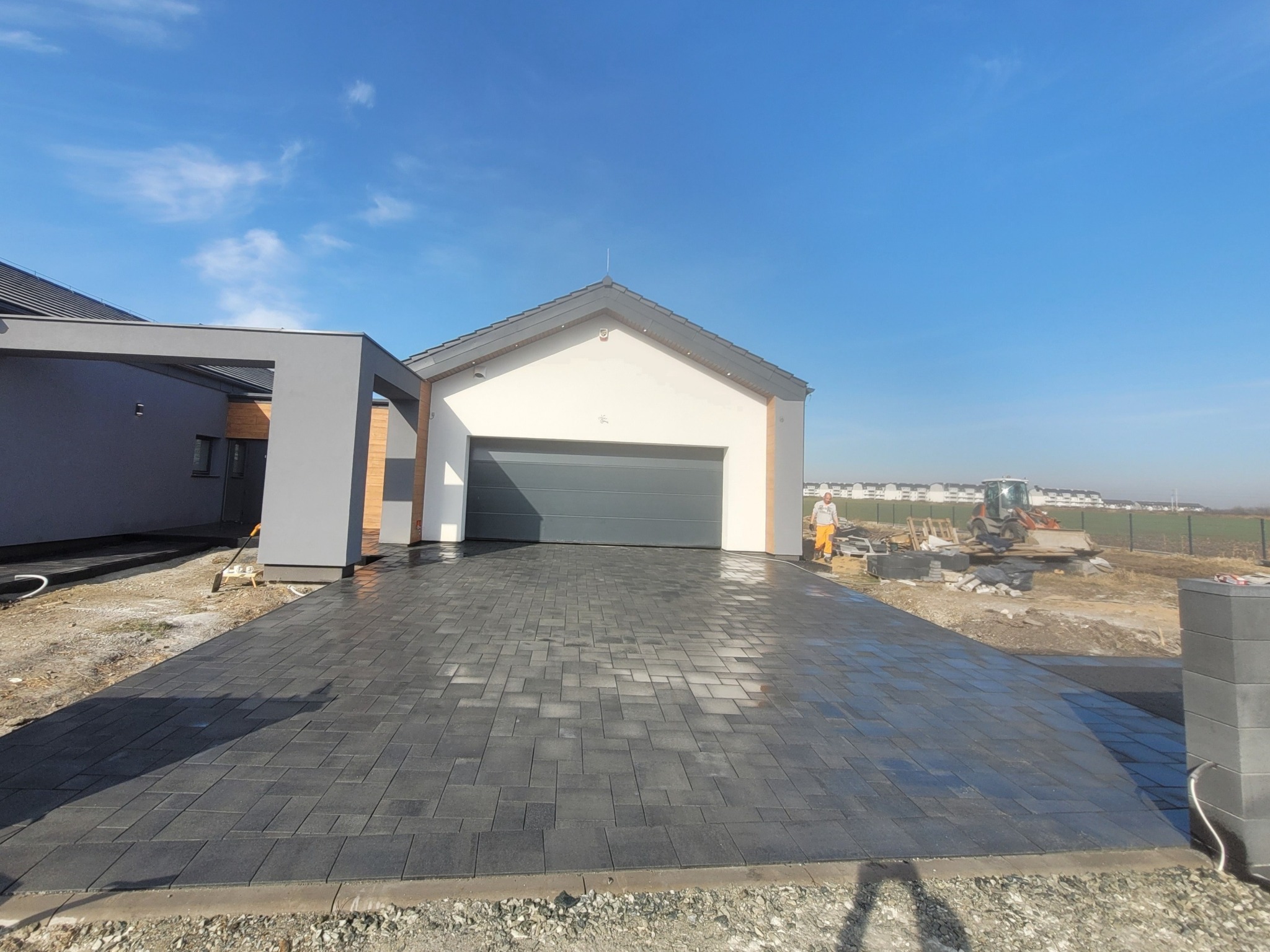 Nowoczesny podjazd z ciemnoszarej kostki betonowej prowadzący do garażu. W tle widać pracownika i sprzęt budowlany, co sugeruje trwające prace wykończeniowe.