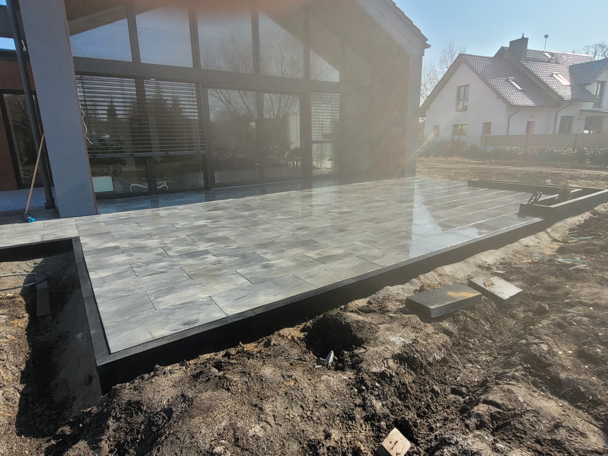 Nowoczesny taras z szarej kostki brukowej, czarne obrzeża, w tle dom z dużymi oknami. Częściowo ukończony, widoczna ziemia. Słoneczny dzień.