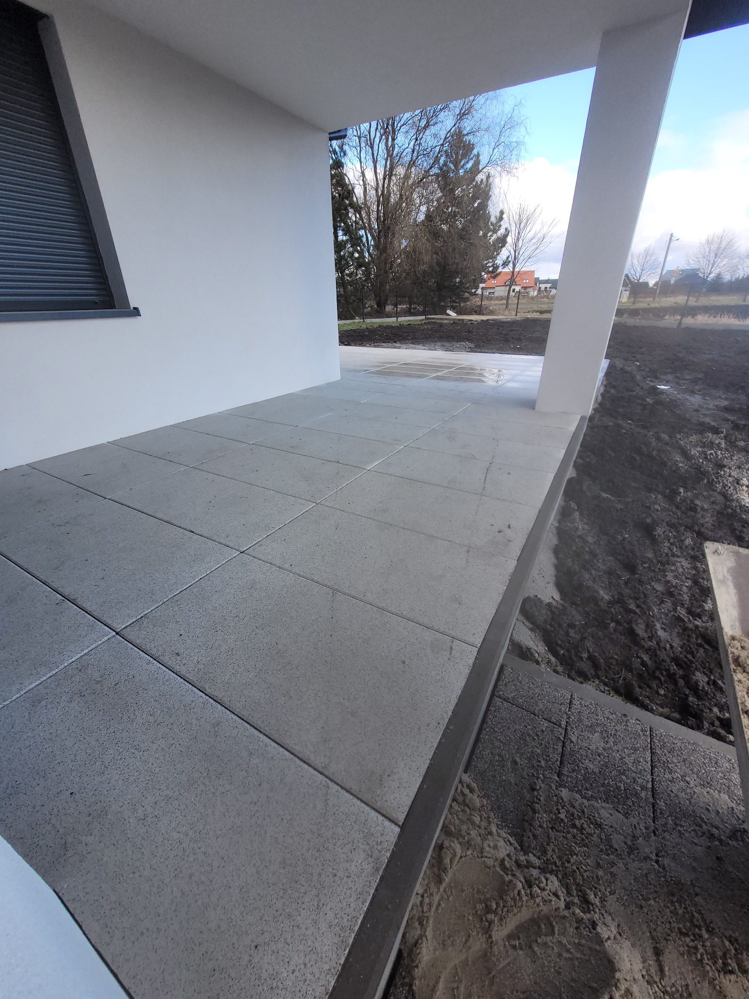 Nowoczesny taras z szarych płyt betonowych, zarys budynku w tle, ziemia przygotowana pod nasadzenia. Minimalistyczny design z widocznymi detalami wykończenia.