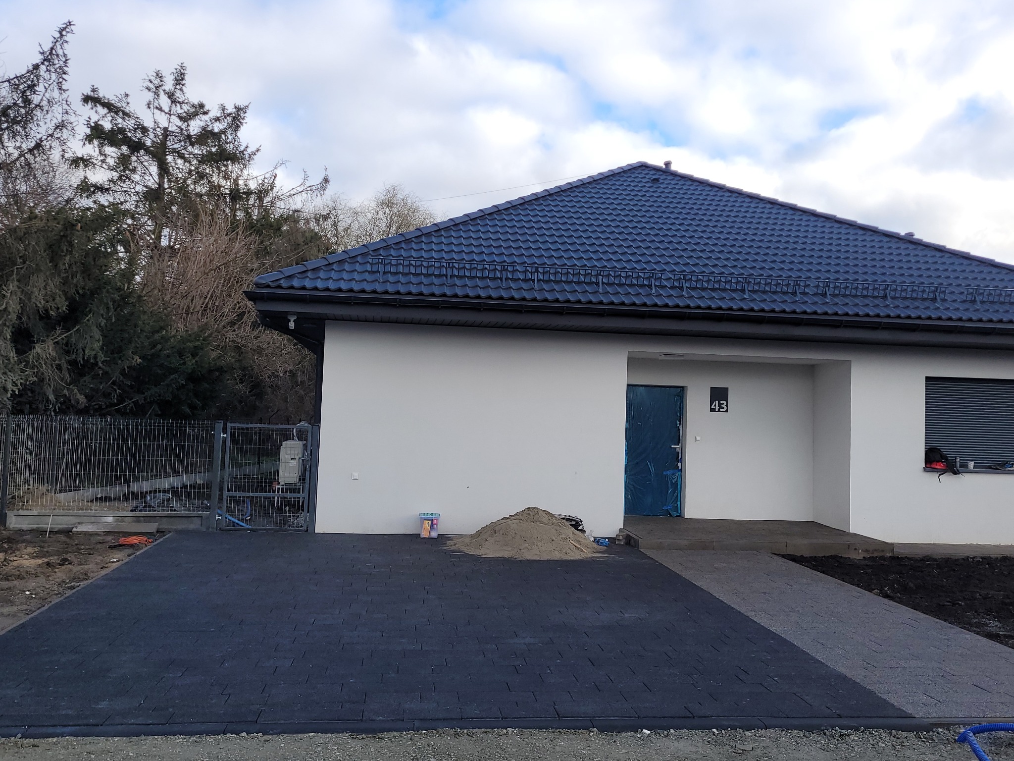 Nowoczesny dom z ciemną kostką brukową na podjeździe. Widoczny fragment ogrodzenia i sterta piasku przed wejściem. Dom ma niebieski dach i numer 43 nad drzwiami.