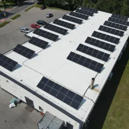 Widok z góry na biały budynek z zamontowanymi panelami słonecznymi, obok parking z zaparkowanymi samochodami i drzewa.