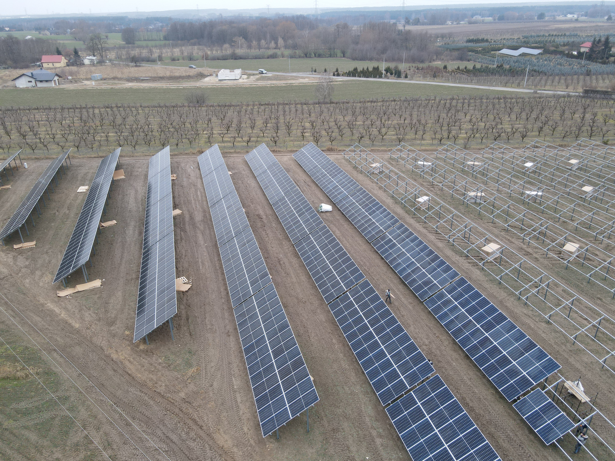 Widok z drona na częściowo ukończoną farmę paneli słonecznych na polu, z widocznymi konstrukcjami wsporczymi i pracującym mężczyzną w oddali.