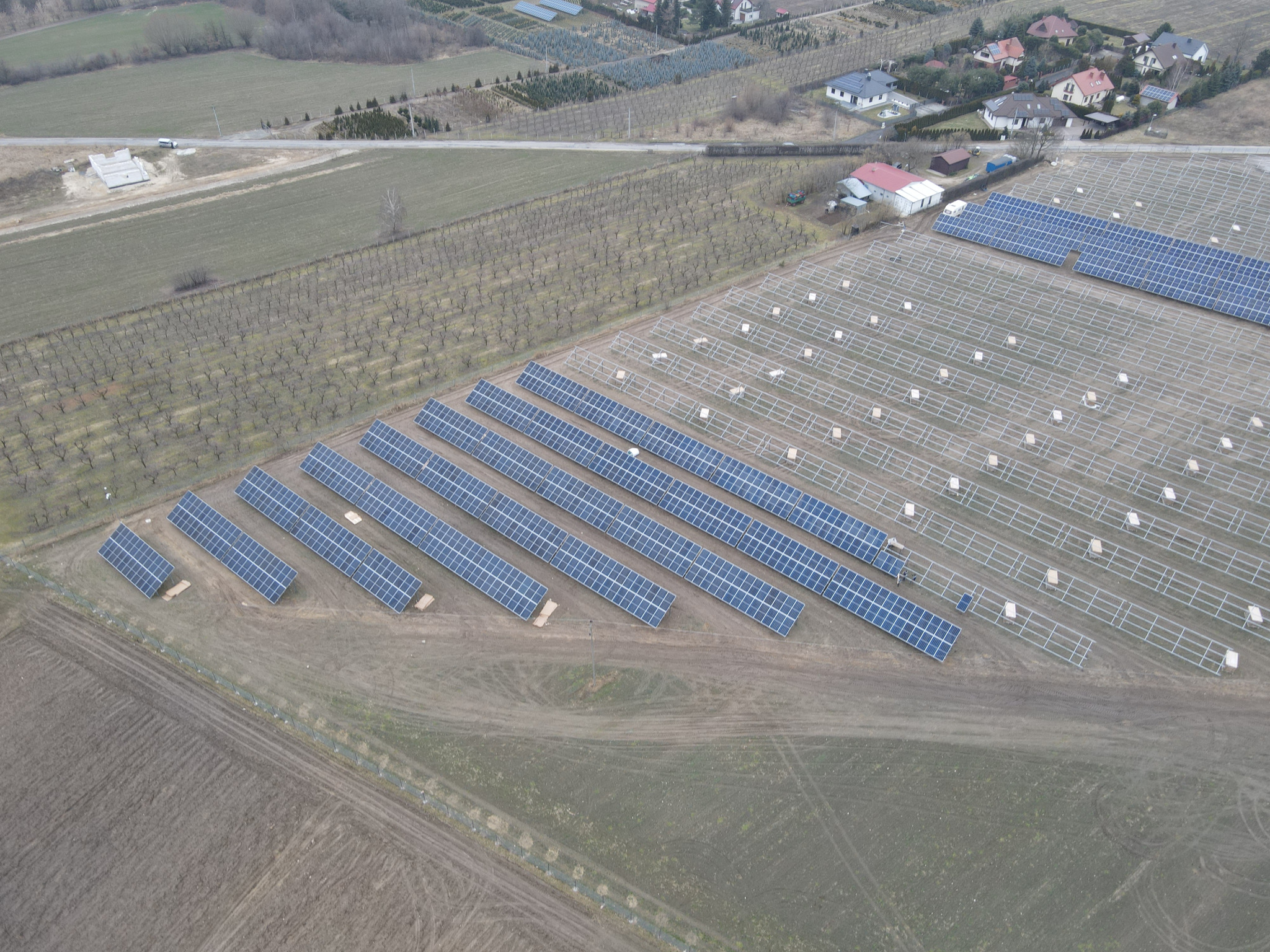 Widok z lotu ptaka na częściowo zainstalowaną farmę paneli słonecznych na polu, z widocznymi rzędami paneli i konstrukcjami wsporczymi, w tle drzewa i zabudowania.