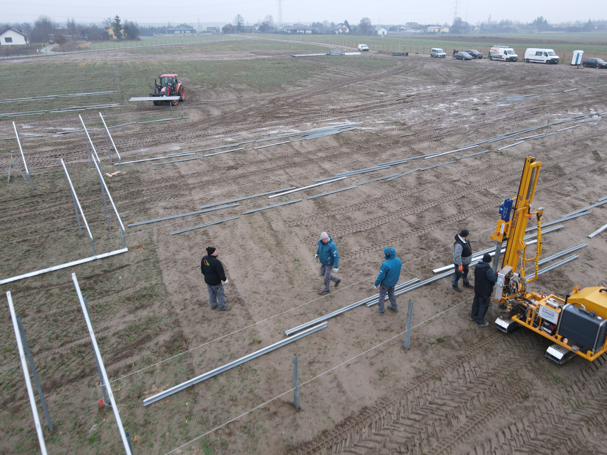 Montaż konstrukcji pod panele słoneczne na błotnistym polu, widoczny żółty sprzęt wiertniczy i grupa monterów w roboczych ubraniach, w tle zabudowania i traktor.