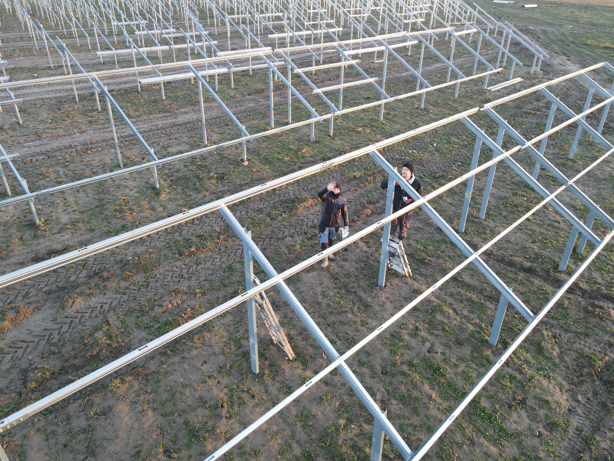 Montaż stelażu pod panele słoneczne na polu, widok z góry na dwóch instalatorów pracujących na drabinie przy skręcaniu konstrukcji z profili aluminiowych.