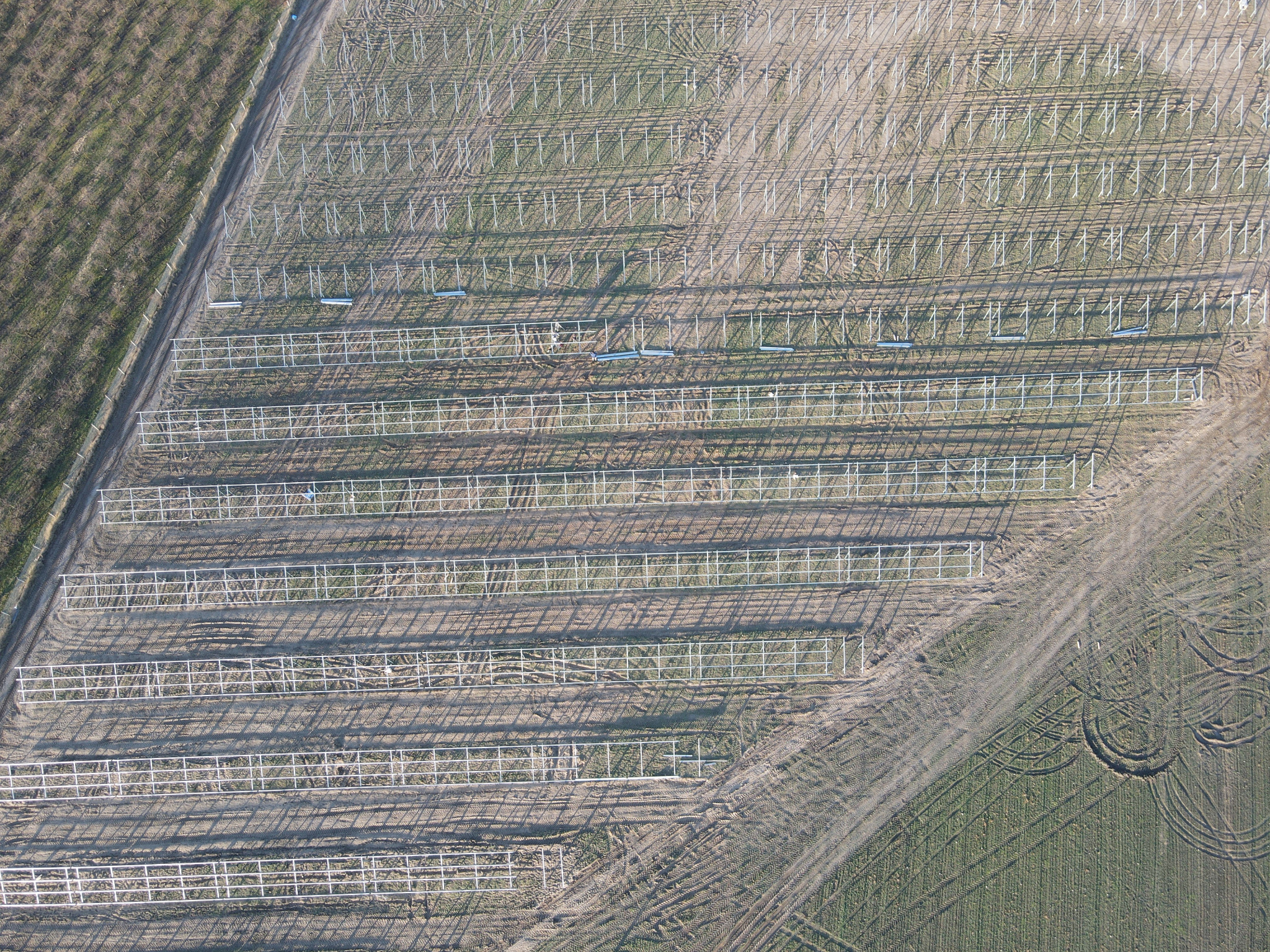 Widok z góry na konstrukcję wsporczą pod panele słoneczne, z widocznymi rzędami metalowych ram i słupków na przygotowanym terenie, częściowo porośniętym trawą, z wyraźnymi śladami opon.