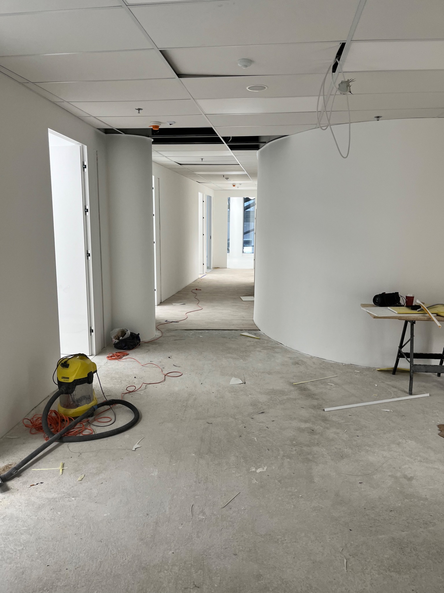 Wnętrze biurowe w trakcie prac wykończeniowych, z widocznym odkurzaczem przemysłowym, niedokończonymi ścianami i sufitem podwieszanym, oraz stolikiem roboczym z narzędziami.