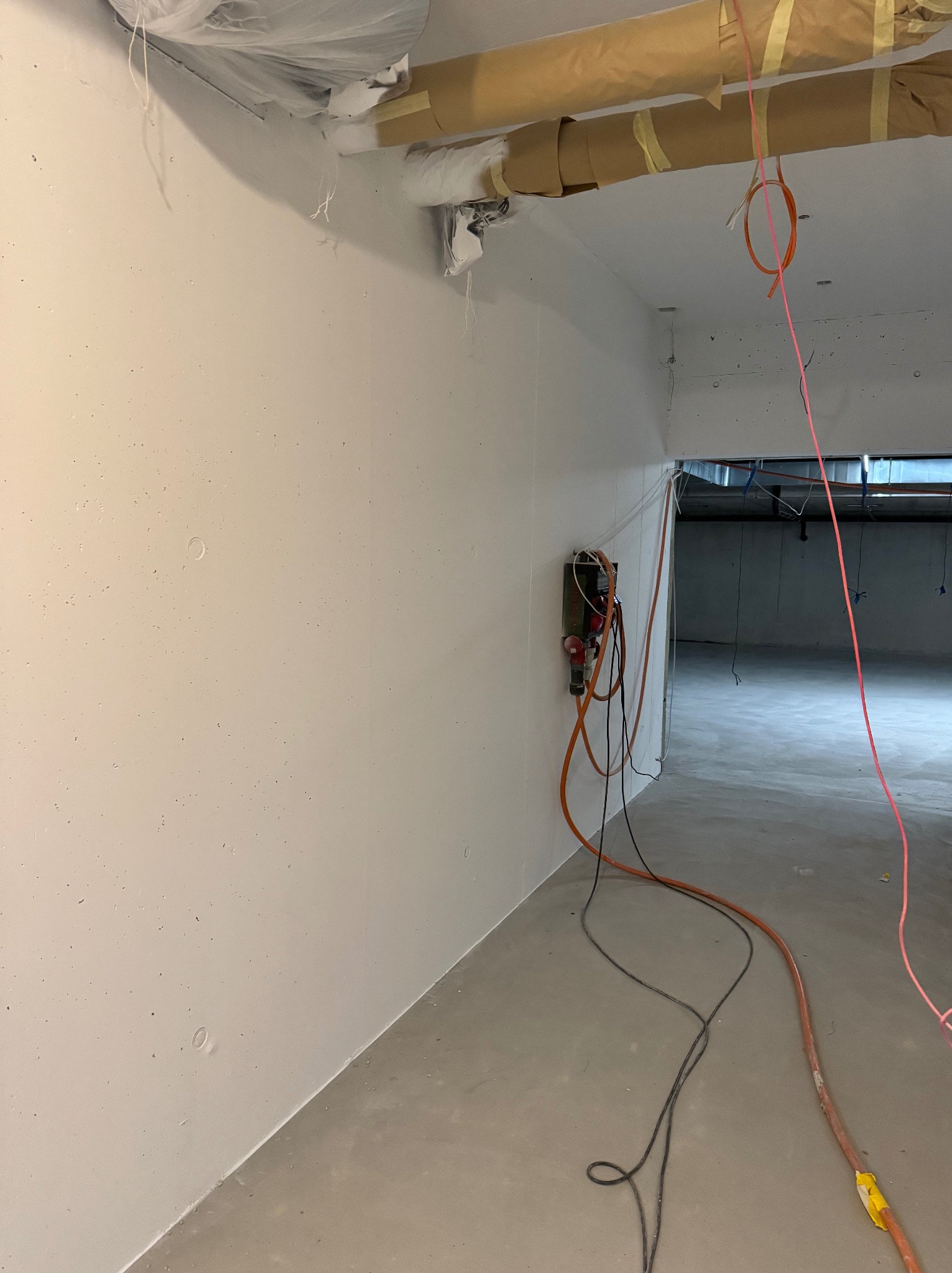 Wnętrze w trakcie prac wykończeniowych: świeżo pomalowana ściana w jasnym odcieniu z widocznymi drobnymi niedoskonałościami, zabezpieczone rury pod sufitem taśmą malarską, wystające kable...