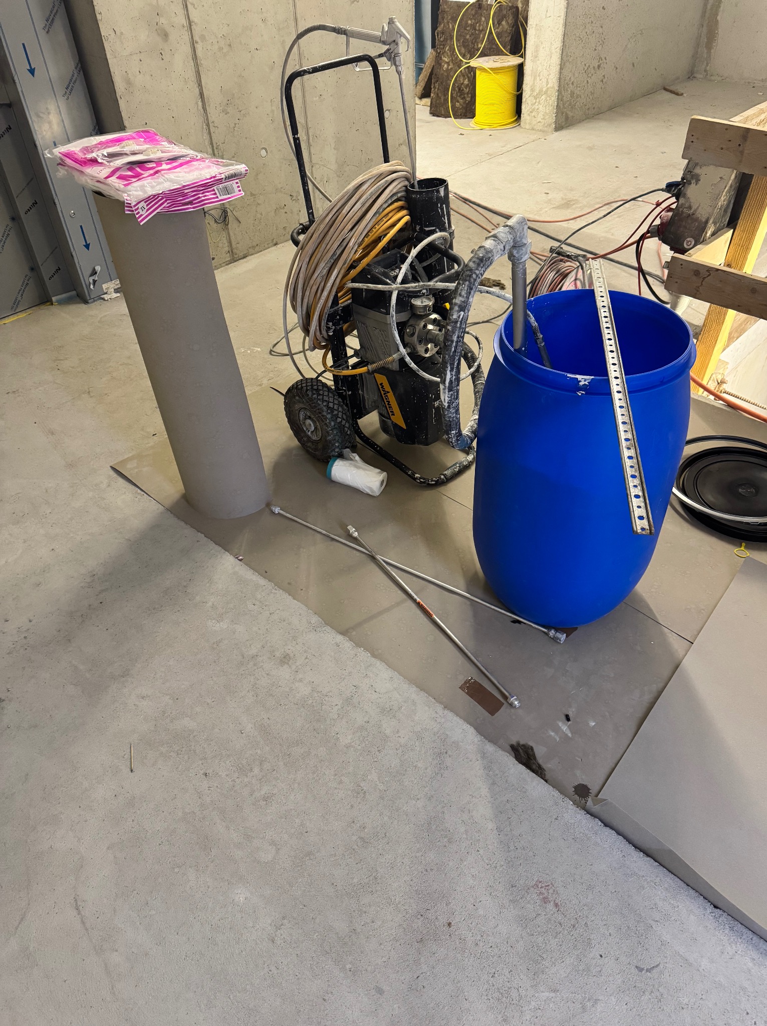 Sprzęt do malowania natryskowego z niebieskim pojemnikiem na farbę, rurami i akcesoriami w surowym wnętrzu, widoczne zabezpieczenia podłoża.