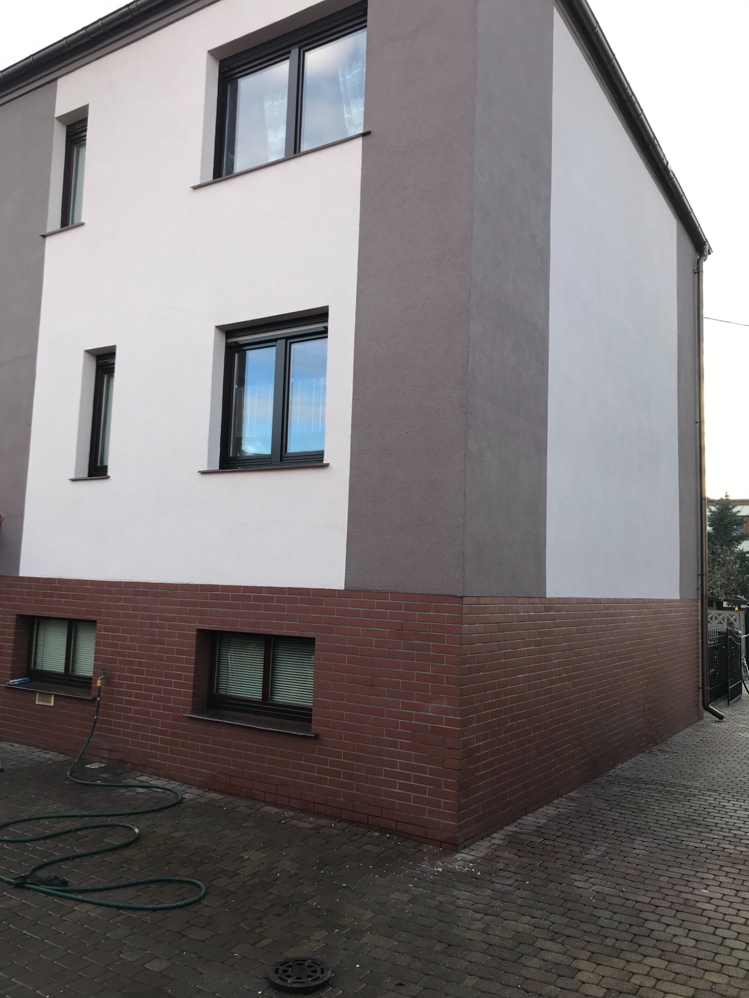 Dwukolorowa elewacja budynku z ceglaną podstawą, widoczne okna z ciemnymi ramami i zielony wąż ogrodowy na brukowanym chodniku.