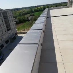 Widok z góry na płaski dach z szarymi panelami obróbki blacharskiej i jasnymi płytkami, w tle budynek w stanie surowym i krajobraz z polami.