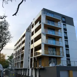 Nowoczesny budynek apartamentowy z balkonami zabezpieczonymi szklanymi balustradami, widok z dołu na elewację w trakcie wykończenia terenu.