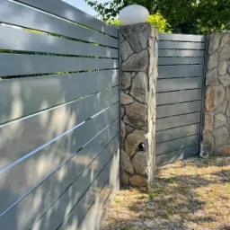 Nowoczesna brama wjazdowa z szarych, poziomych paneli, osadzona na kamiennych słupkach zwieńczonych kulistymi lampami, na tle zieleni ogrodu.