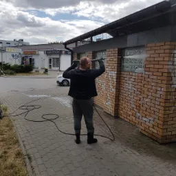 Mężczyzna w okularach ochronnych czyści ceglaną ścianę budynku z graffiti za pomocą myjki ciśnieniowej, widoczna maszyna czyszcząca, tło stanowią budynki usługowe.