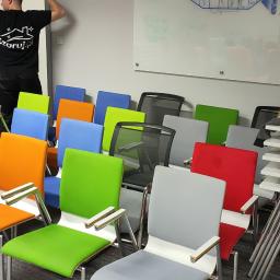 SZORUJ.PL SPÓŁKA Z OGRANICZONĄ ODPOWIEDZIALNOŚCIĄ - Rząd kolorowych krzeseł biurowych z chromowanymi podłokietnikami w sali konferencyjnej, za nimi biała tablica i pracownik w czarnej koszulce z logo firmy sprzątającej.