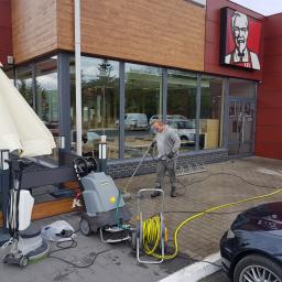 SZORUJ.PL SPÓŁKA Z OGRANICZONĄ ODPOWIEDZIALNOŚCIĄ - Pracownik w okularach ochronnych czyści kostkę brukową przed restauracją KFC za pomocą myjki ciśnieniowej Kärcher. Na pierwszym planie zwinięty żółty wąż, obok maszyna do czyszczenia podłóg.