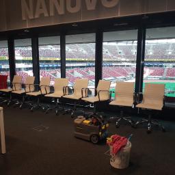 SZORUJ.PL SPÓŁKA Z OGRANICZONĄ ODPOWIEDZIALNOŚCIĄ - Rząd białych krzeseł biurowych ustawionych wzdłuż okna z widokiem na stadion, obok stoi odkurzacz piorący Karcher i wiadro z mopami.