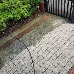 SZORUJ.PL SPÓŁKA Z OGRANICZONĄ ODPOWIEDZIALNOŚCIĄ - Częściowo umyta kostka brukowa w odcieniach szarości i brązu, widoczny wąż ciśnieniowy i brama z czarnego metalu na tle zieleni.