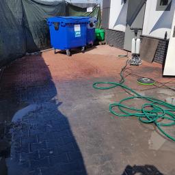 SZORUJ.PL SPÓŁKA Z OGRANICZONĄ ODPOWIEDZIALNOŚCIĄ - Mokre, czerwono-czarne kostki brukowe po czyszczeniu, widoczne dwa niebieskie kontenery na kółkach, zwinięty zielony wąż ogrodowy oraz maszyna czyszcząca z padem szorującym.