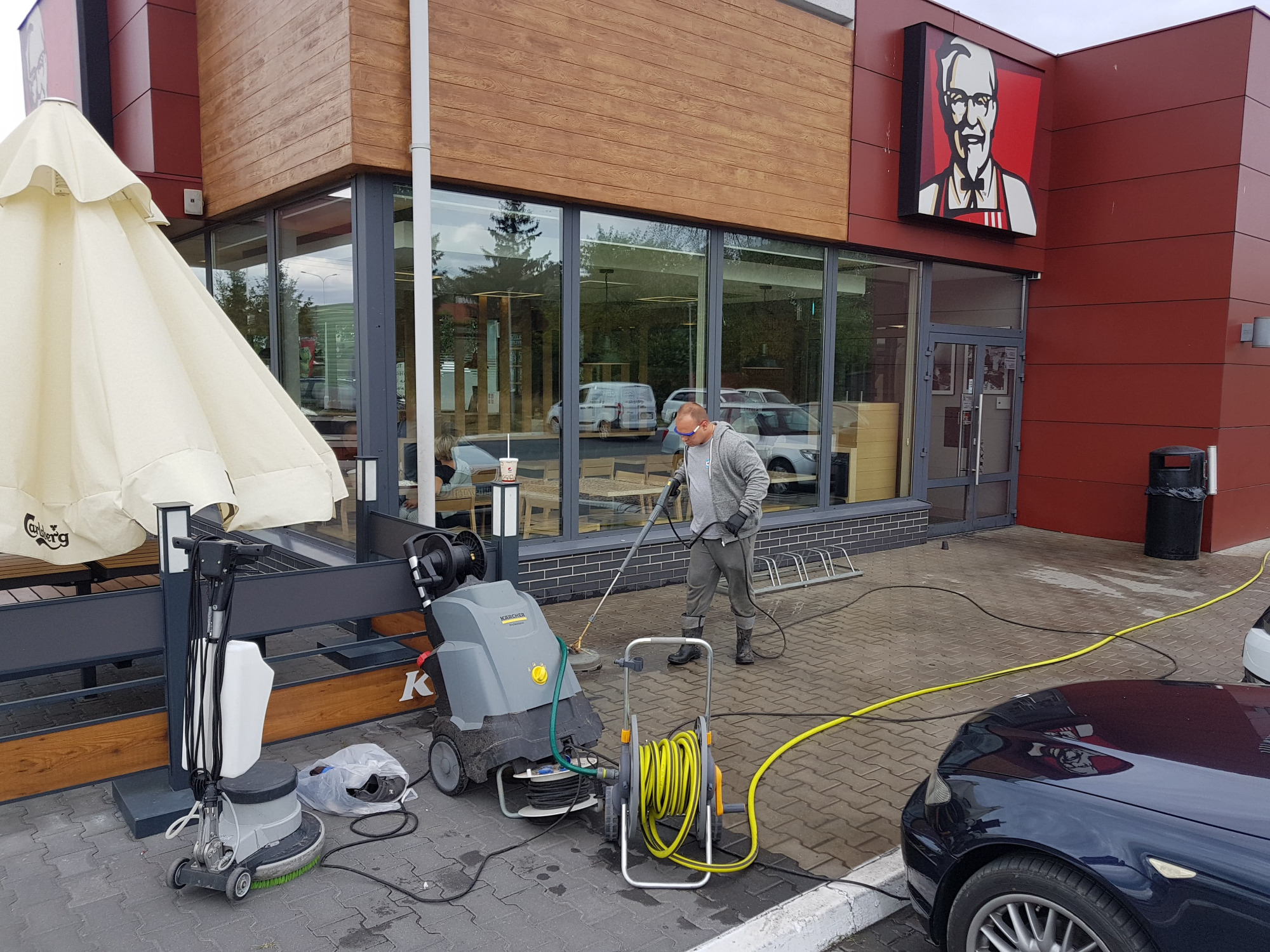 Pracownik w okularach ochronnych czyści kostkę brukową przed restauracją KFC za pomocą myjki ciśnieniowej Kärcher. Na pierwszym planie zwinięty żółty wąż, obok maszyna do czyszczenia podłóg.