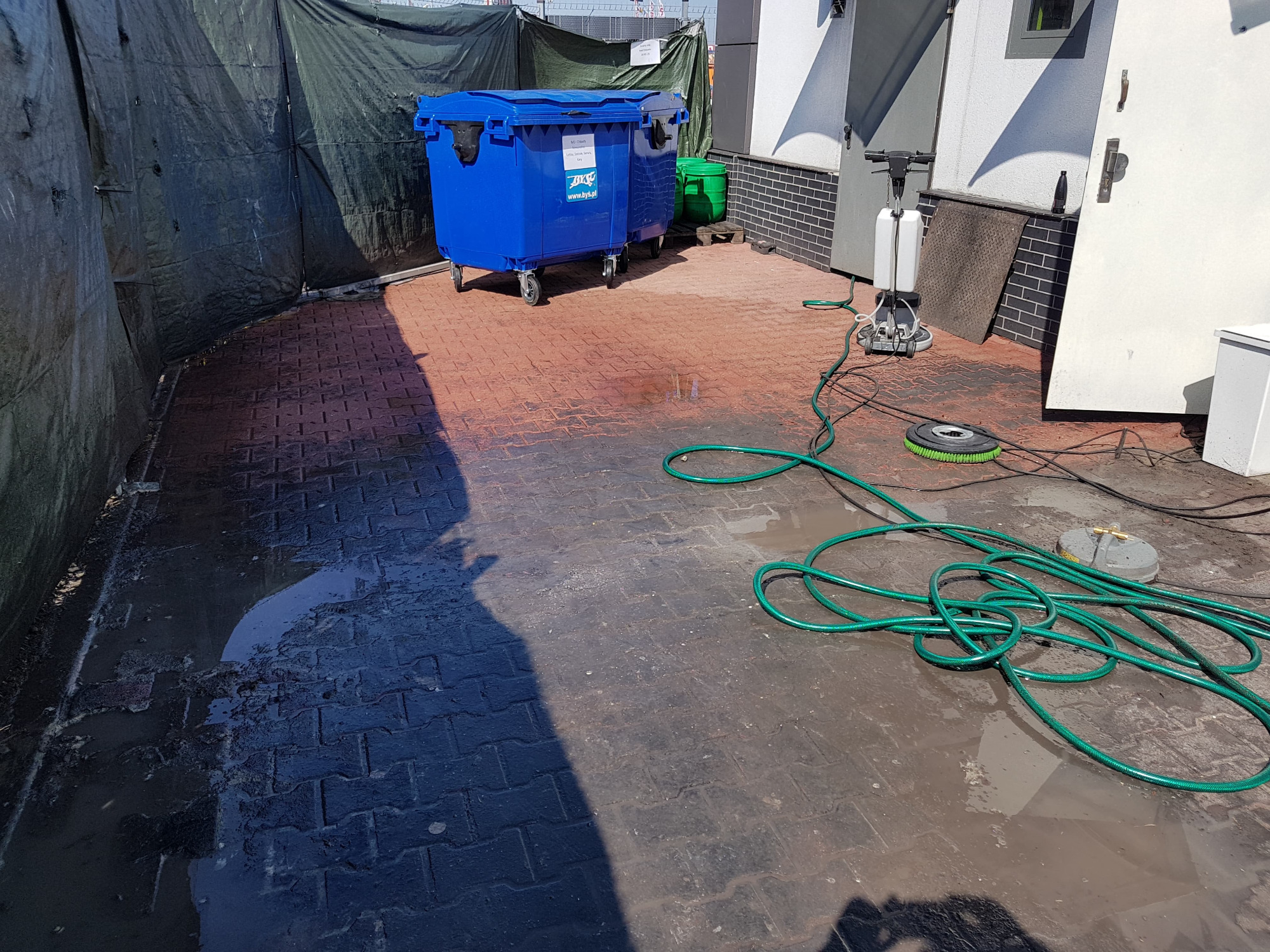 Mokre, czerwono-czarne kostki brukowe po czyszczeniu, widoczne dwa niebieskie kontenery na kółkach, zwinięty zielony wąż ogrodowy oraz maszyna czyszcząca z padem szorującym.