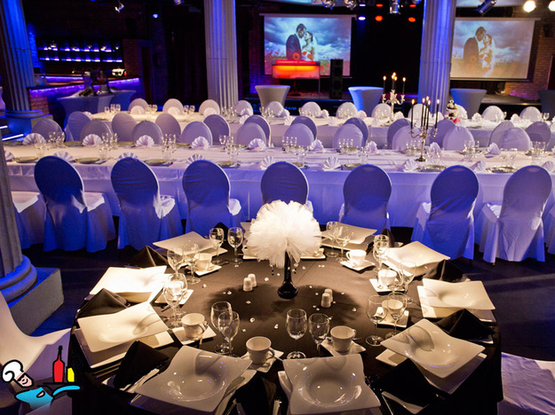 Uroczyste nakrycie stołu z czarnym obrusem, białą dekoracją z piór, kwadratową zastawą i kryształowymi kieliszkami, w tle sala balowa z kolumnami i projekcją wideo.