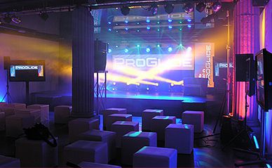 Sala eventowa oświetlona reflektorami, z niską sceną, kolumną architektoniczną i białymi pufami dla gości.