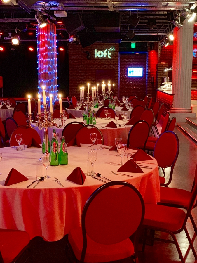 Uroczysta sala z okrągłymi stołami nakrytymi białymi obrusami, czerwonymi serwetkami i krzesłami, świeczniki ze świecami, butelki wody mineralnej, w tle kolumna z oświetleniem LED i logo Loft44...