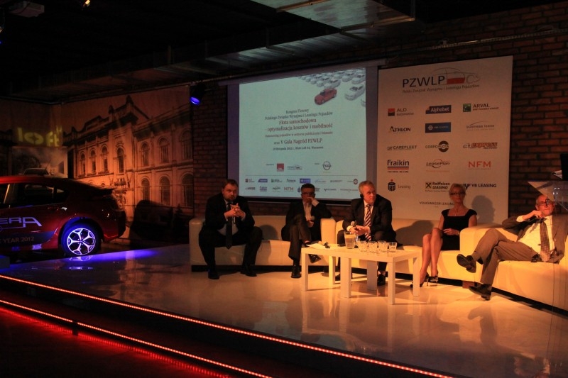 Panel dyskusyjny na scenie z czerwonym samochodem elektrycznym w tle, logo PZWLP i sponsorów na ścianie, oświetlenie sceniczne.