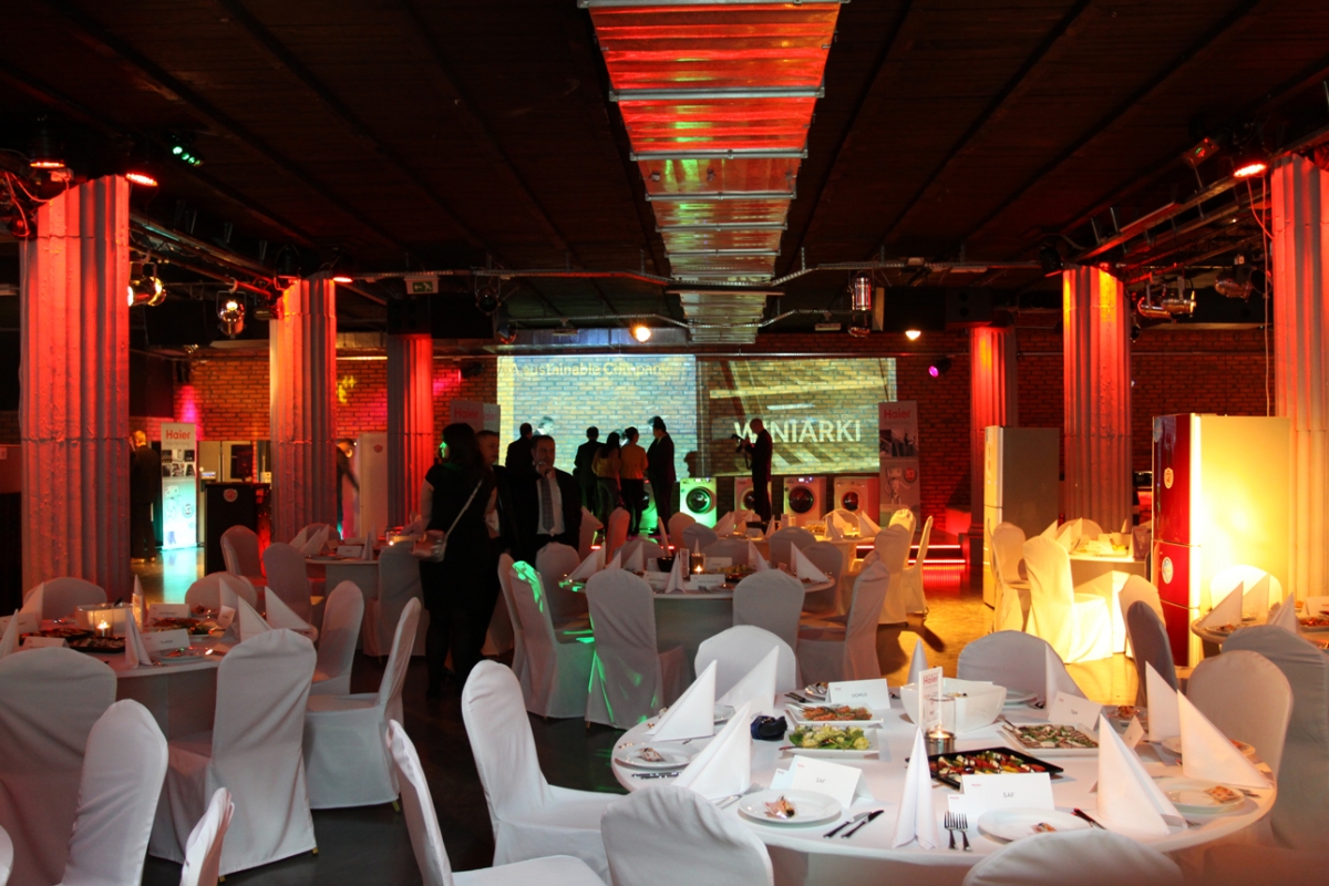 Sala bankietowa z okrągłymi stołami nakrytymi białymi obrusami i krzesłami w pokrowcach, oświetlona czerwonymi i zielonymi światłami, z widocznymi kolumnami i ekranem projekcyjnym w tle.