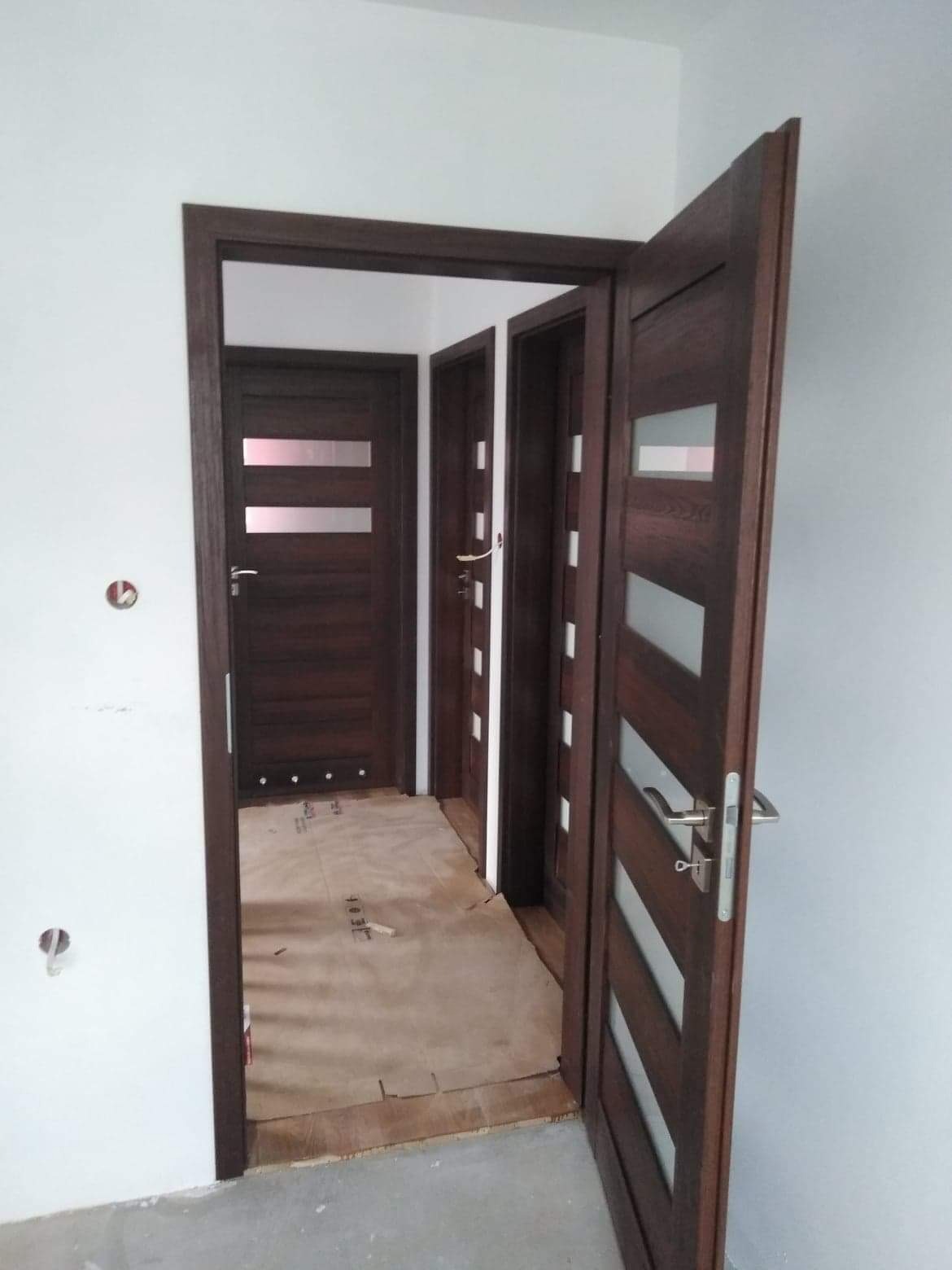 Nowo zamontowane, ciemnobrązowe drzwi wewnętrzne z przeszkleniami, widoczne trzy pary drzwi w perspektywie, podłoga zabezpieczona kartonem.