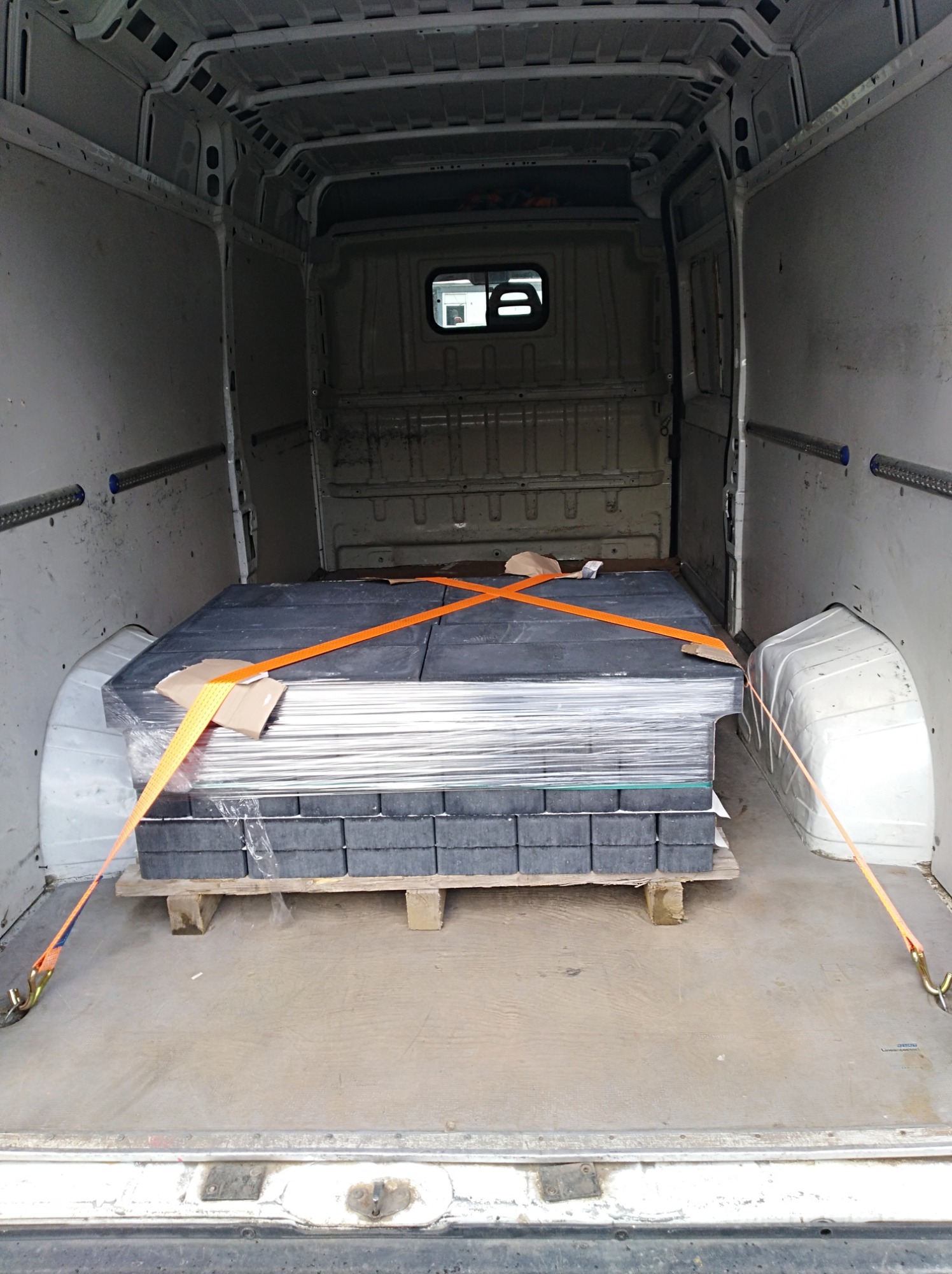 Wnętrze furgonetki z paletą kostki brukowej, zabezpieczoną pomarańczowymi pasami transportowymi, widoczne ślady użytkowania na podłodze.