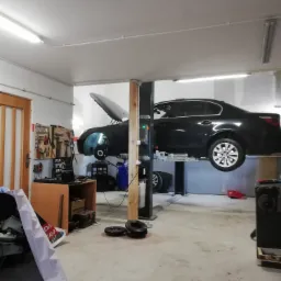 Czarny samochód osobowy na podnośniku dwukolumnowym w warsztacie, z otwartą maską i zdjętym kołem, widoczne narzędzia na ścianie.