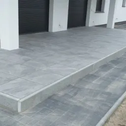 Szare płytki tarasowe ułożone przed domem w Zagórzu, widoczne również schody z płytek i fragment ogrodzenia w tle, na pierwszym planie robocze rękawice.