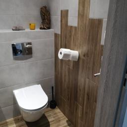 RemontNa5 - Nowoczesna toaleta z szarymi płytkami i drewnopodobnymi akcentami na ścianie, wisząca muszla klozetowa, drewniana podłoga i dekoracje na półce