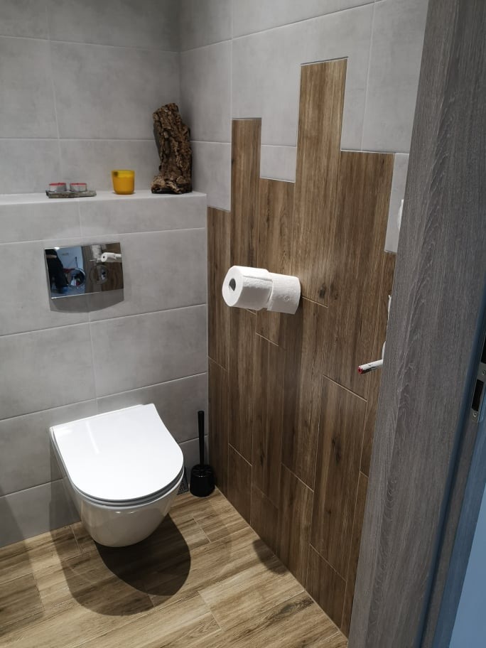 Nowoczesna toaleta z szarymi płytkami i drewnopodobnymi akcentami na ścianie, wisząca muszla klozetowa, drewniana podłoga i dekoracje na półce