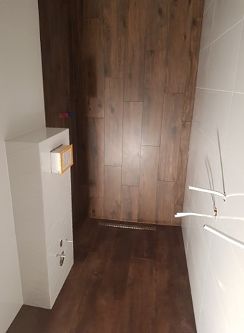 Wykończona łazienka z podłogą i ścianą prysznicową wyłożoną płytkami imitującymi ciemne drewno, odpływ liniowy w podłodze, jasne płytki na pozostałych ścianach, widoczne rury i elementy instalacji.