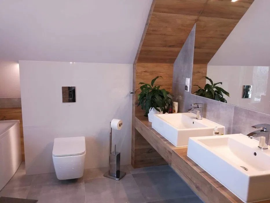 Nowoczesna łazienka na poddaszu z dwoma białymi umywalkami nablatowymi, drewnianym blatem i lustrem, widoczna toaleta podwieszana z chromowanym przyciskiem spłukiwania i stojak na papier toaletowy.
