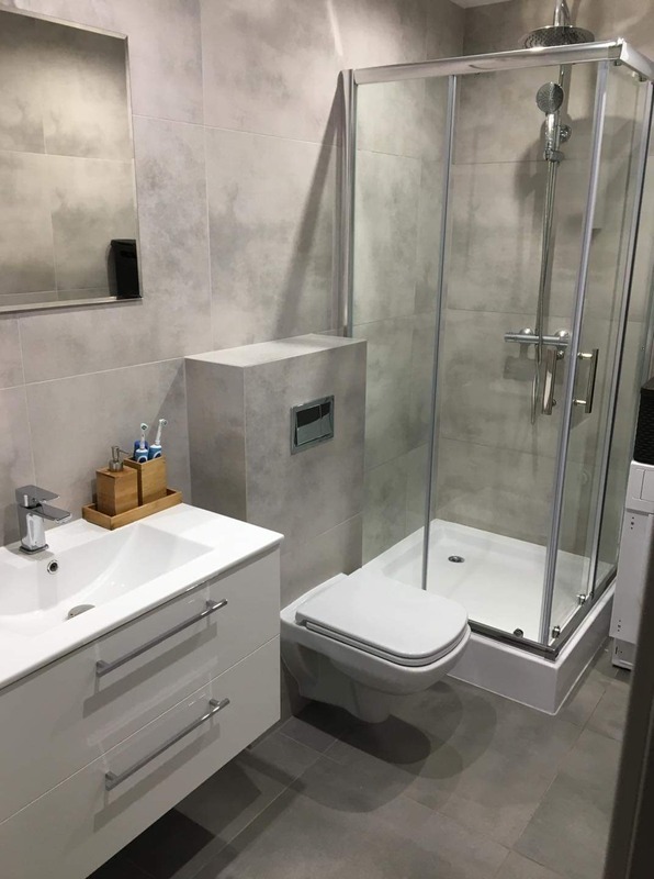 Nowoczesna łazienka z kabiną prysznicową, umywalką z szafką i podwieszaną toaletą, wykończona szarymi płytkami imitującymi beton.